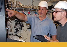 A thumbnail image of two people in hard hats in an environment with many wires. One person is pointing at something and the other person is taking.